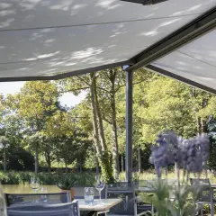 Terraza con mesas y sillas bajo una lona que es un toldo retráctil, con vistas a un río y árboles frondosos.