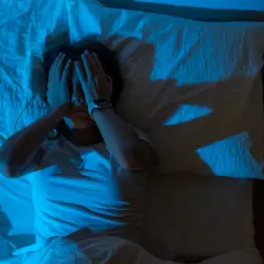 Persona en la cama, con las manos cubriendo su rostro, en un ambiente oscuro y con luz azul. Parece estar despertándose de golpe asustado.