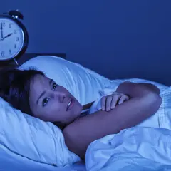 Mujer en la cama, con los ojos muy abiertos, sintiendo que se está despertando de golpe con taquicardia. Un despertador antiguo marca las 3:10 AM.