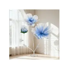 Tres flores de tela grandes, azules y blancas, con tallos blancos, junto a una ventana con cortinas.