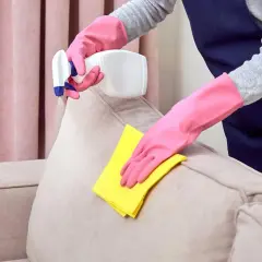 Manos enguantadas limpian un sofá beige con un paño amarillo y spray.
