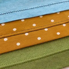 Demostración de como hacer un dobladillo en tela de punto con diferentes puntadas y telas: azul, mostaza con lunares blancos y verde.