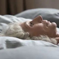 Mujer mayor con cabello blanco yace en la cama, con los ojos entreabiertos, como si soñara que duermo con los ojos abiertos.