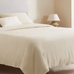 Cama vestida con ropa de cama de lino color crema, que incluye una bajera de cama, almohadas y un edredón.