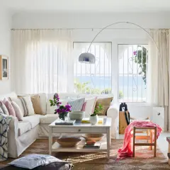 Sofá blanco con cojines de **fibras de algodón** y manta a cuadros, junto a una mesa de centro y ventana con vistas al mar.