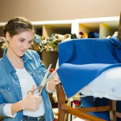 Mujer cortando tela azul para como tapizar un sillón pequeño. Estanterías con rollos de tela al fondo.
