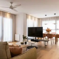 Acogedor salón de 20 metros cuadrados con sofá, mesa de centro, TV, y comedor. Cortinas blancas y suelo de madera crean un ambiente luminoso.