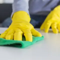 Manos con guantes amarillos limpiando una superficie blanca con un trapo verde. Aprende como lavar los trapos de limpieza para un hogar impecable.