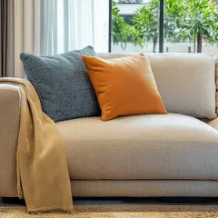 Sofá de tela beige con cojines azul y naranja. Un chal color camel cuelga del brazo. Ideal para aprender como limpiar sofa de tela.