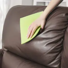 Mano limpiando un sofá de piel marrón con un paño verde. Aprende cómo limpiar sofá de piel para mantenerlo como nuevo.