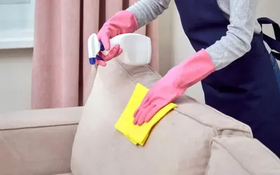 Manos enguantadas limpian un sofá beige con un paño amarillo y spray.