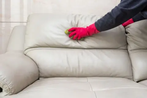 Mano con guante rosa limpiando un sofá de cuero beige con una esponja. Aprende cómo limpiar sofá de cuero para mantenerlo como nuevo.