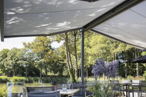 Terraza con mesas y sillas bajo una lona que es un toldo retráctil, con vistas a un río y árboles frondosos.