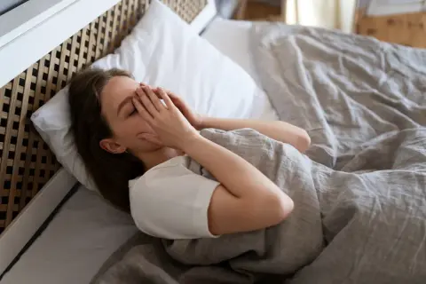 Mujer en la cama, con las manos cubriendo su rostro, luchando contra el miedo a quedarse dormido.