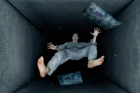 Un hombre en pijama experimenta una caida al vacio dentro de un túnel oscuro, con almohadas flotando a su alrededor.