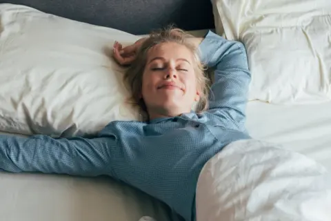 Mujer rubia sonríe plácidamente en su cama, disfrutando de un sueño reparador bajo la suave roba de llit.