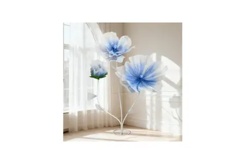 Tres flores de tela grandes, azules y blancas, con tallos blancos, junto a una ventana con cortinas.