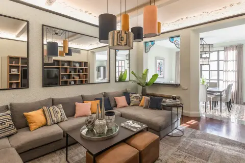 Amplios salones con sofás modulares grises, cojines coloridos y una mesa de centro. Reflejos en espejos y lámparas colgantes añaden estilo.