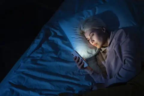 Mujer rubia en la cama, mirando su móvil con luz azul. Quizás por eso me despierto a las 4 de la mañana.