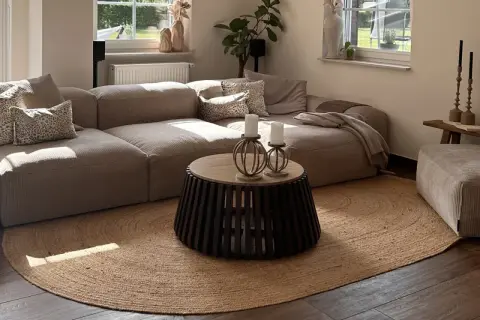 Alfombra redonda de yute planta bajo un sofá modular beige. La luz del sol ilumina la sala.