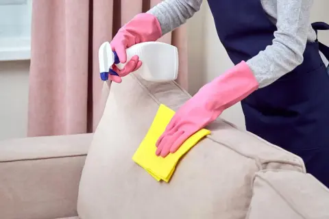 Manos enguantadas limpian un sofá beige con un paño amarillo y spray.