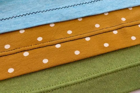 Demostración de como hacer un dobladillo en tela de punto con diferentes puntadas y telas: azul, mostaza con lunares blancos y verde.
