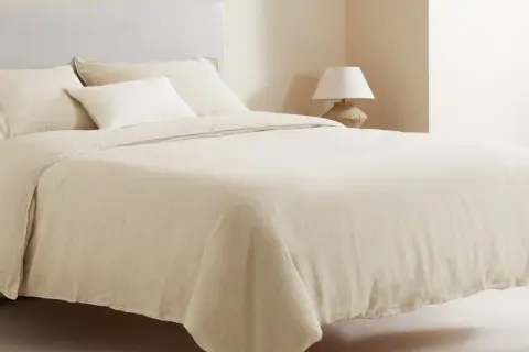 Cama vestida con ropa de cama de lino color crema, que incluye una bajera de cama, almohadas y un edredón.