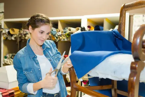Mujer cortando tela azul para como tapizar un sillón pequeño. Estanterías con rollos de tela al fondo.