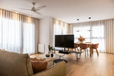 Acogedor salón de 20 metros cuadrados con sofá, mesa de centro, TV, y comedor. Cortinas blancas y suelo de madera crean un ambiente luminoso.