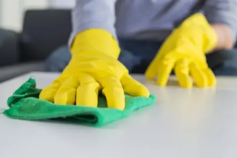 Manos con guantes amarillos limpiando una superficie blanca con un trapo verde. Aprende como lavar los trapos de limpieza para un hogar impecable.