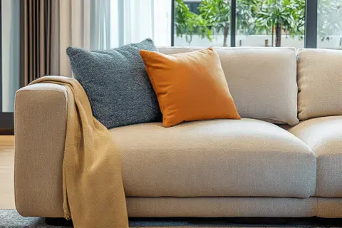 Sofá de tela beige con cojines azul y naranja. Un chal color camel cuelga del brazo. Ideal para aprender como limpiar sofa de tela.