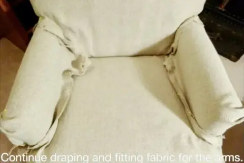 Tela beige drapeada y ajustada con alfileres en los brazos de una silla, parte de un tutorial sobre como hacer fundas para sillas.
