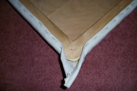 Detalle de cómo tapizar esquinas de un bastidor de madera con tela blanca, asegurada con grapas.