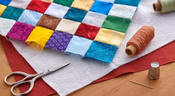 Un trozo de tela de patchwork con cuadrados de colores, tijeras, hilos y dedal. Así se empieza un proyecto de patchwork.