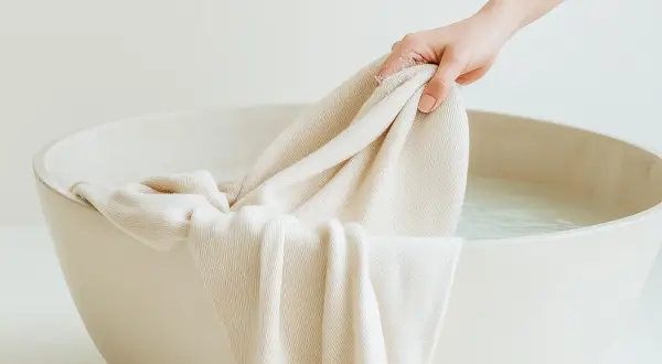 Mano sumerge tela clara en agua para lavar ropa blanca.