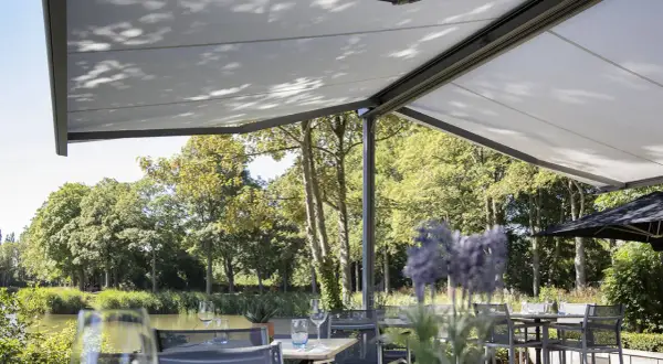 Terraza con mesas y sillas bajo una lona que es un toldo retráctil, con vistas a un río y árboles frondosos.