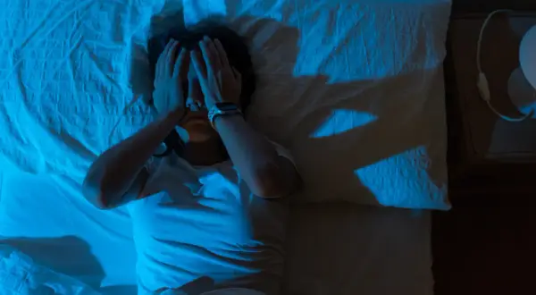 Persona en la cama, con las manos cubriendo su rostro, en un ambiente oscuro y con luz azul. Parece estar despertándose de golpe asustado.