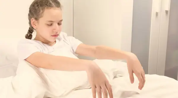 Niña con trenzas, ojos cerrados, sentada en la cama con los brazos caídos, como una persona sonámbula.