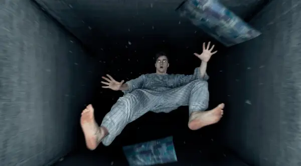 Un hombre en pijama experimenta una caida al vacio dentro de un túnel oscuro, con almohadas flotando a su alrededor.