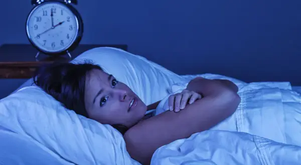 Mujer en la cama, con los ojos muy abiertos, sintiendo que se está despertando de golpe con taquicardia. Un despertador antiguo marca las 3:10 AM.
