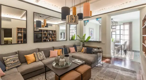 Amplios salones con sofás modulares grises, cojines coloridos y una mesa de centro. Reflejos en espejos y lámparas colgantes añaden estilo.