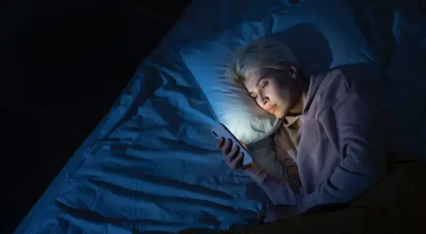 Mujer rubia en la cama, mirando su móvil con luz azul. Quizás por eso me despierto a las 4 de la mañana.