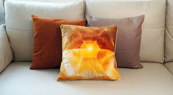 Tres cojines sobre un sofá de cuero: uno naranja texturizado, uno con un diseño de cristal amarillo brillante y otro marrón. El cojín central, con su estampado de velour que es, irradia calidez.