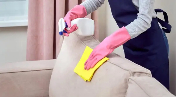 Manos enguantadas limpian un sofá beige con un paño amarillo y spray.