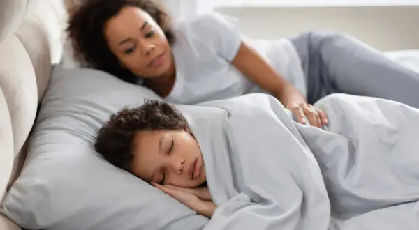 Madre observa a su hijo dormir plácidamente. Quizás se pregunta por qué se habla en sueños, o simplemente disfruta de la paz del momento.