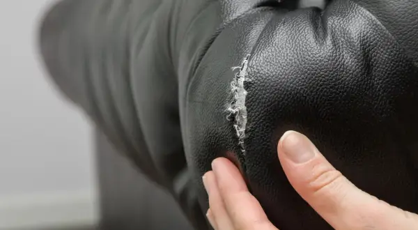 Mano examinando un desgarro en cuero negro, mostrando la necesidad de reparar cuero agrietado.