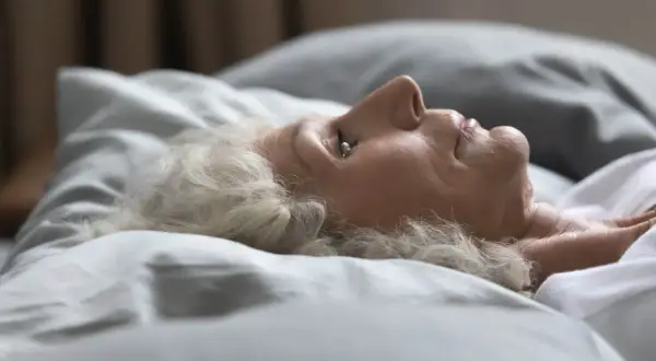 Mujer mayor con cabello blanco yace en la cama, con los ojos entreabiertos, como si soñara que duermo con los ojos abiertos.
