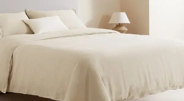 Cama vestida con ropa de cama de lino color crema, que incluye una bajera de cama, almohadas y un edredón.