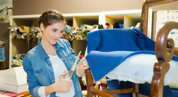 Mujer cortando tela azul para como tapizar un sillón pequeño. Estanterías con rollos de tela al fondo.