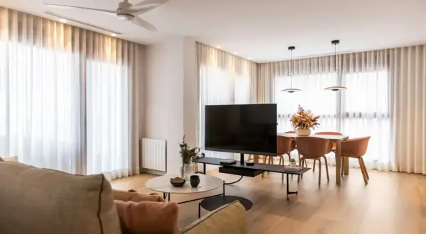 Acogedor salón de 20 metros cuadrados con sofá, mesa de centro, TV, y comedor. Cortinas blancas y suelo de madera crean un ambiente luminoso.